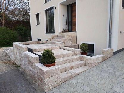 Blockstufen aus Travertin Classic Beige als breite Außentreppe mit Podest und seitlichen Natursteinmauern vor einem Wohnhaus.